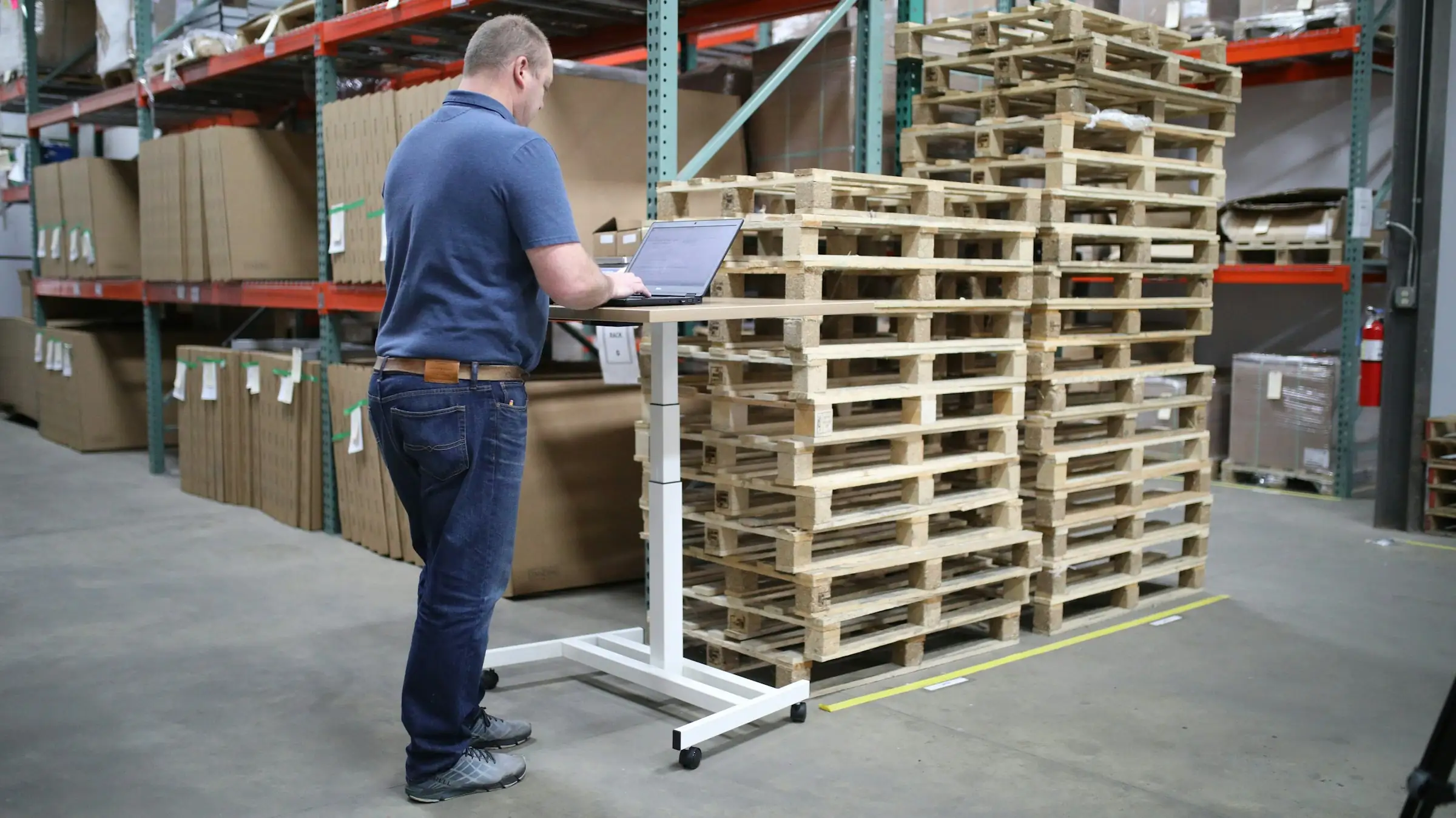 Warehouse manager working on a laptop at a mobile standing desk beside pallets